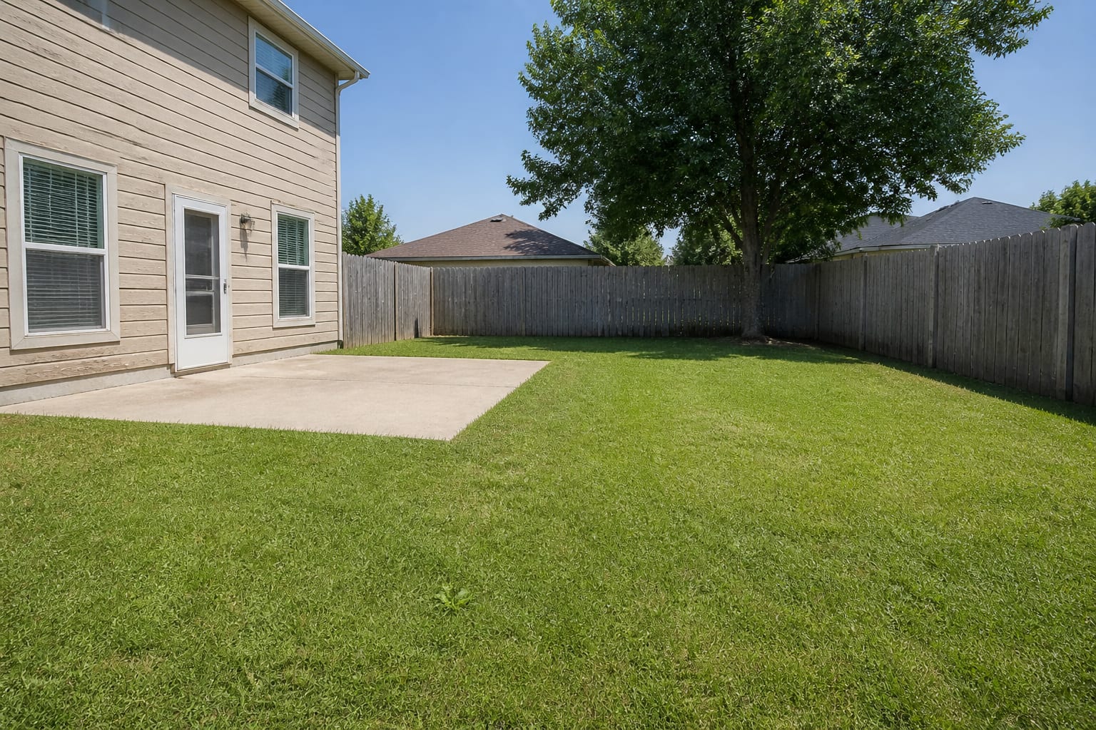 A plain suburban backyard with a flat green lawn and an empty gray concrete patio before redesign