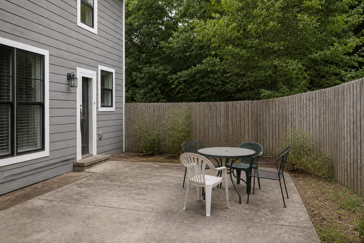 Narrow patio corner with a small metal table, mismatched chairs, no shade, and bare concrete around the dining area.