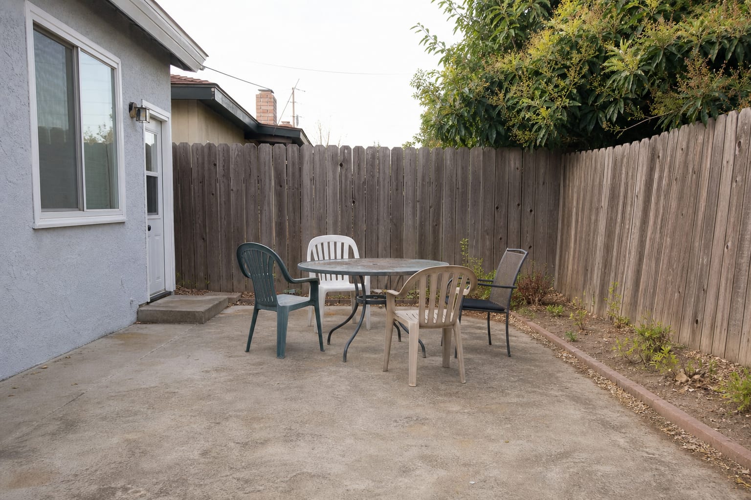 Narrow patio corner with a small metal table, mismatched chairs, no shade, and bare concrete around the dining area.