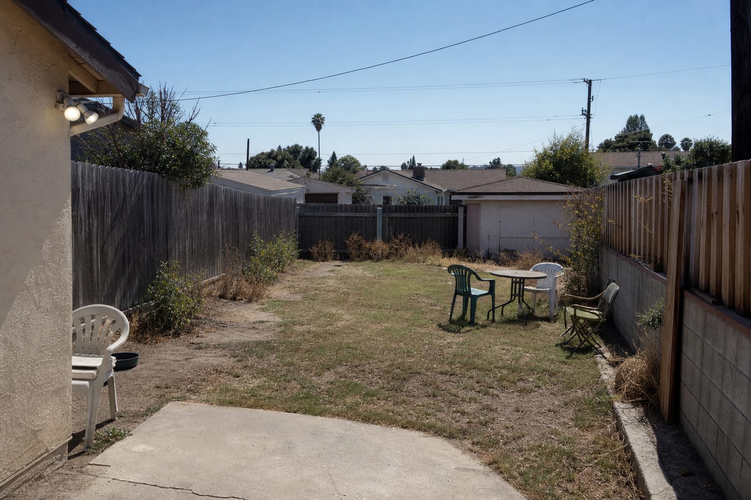 underused backyard with scattered chairs, bare grass, no shade, a small patio table, and one bright wall light near the back door.