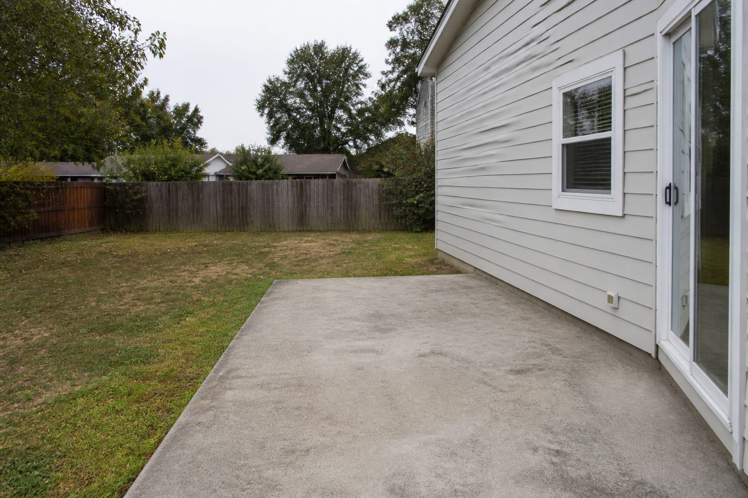 Plain backyard with empty concrete pad and overgrown grass — no fitness equipment or defined training zone