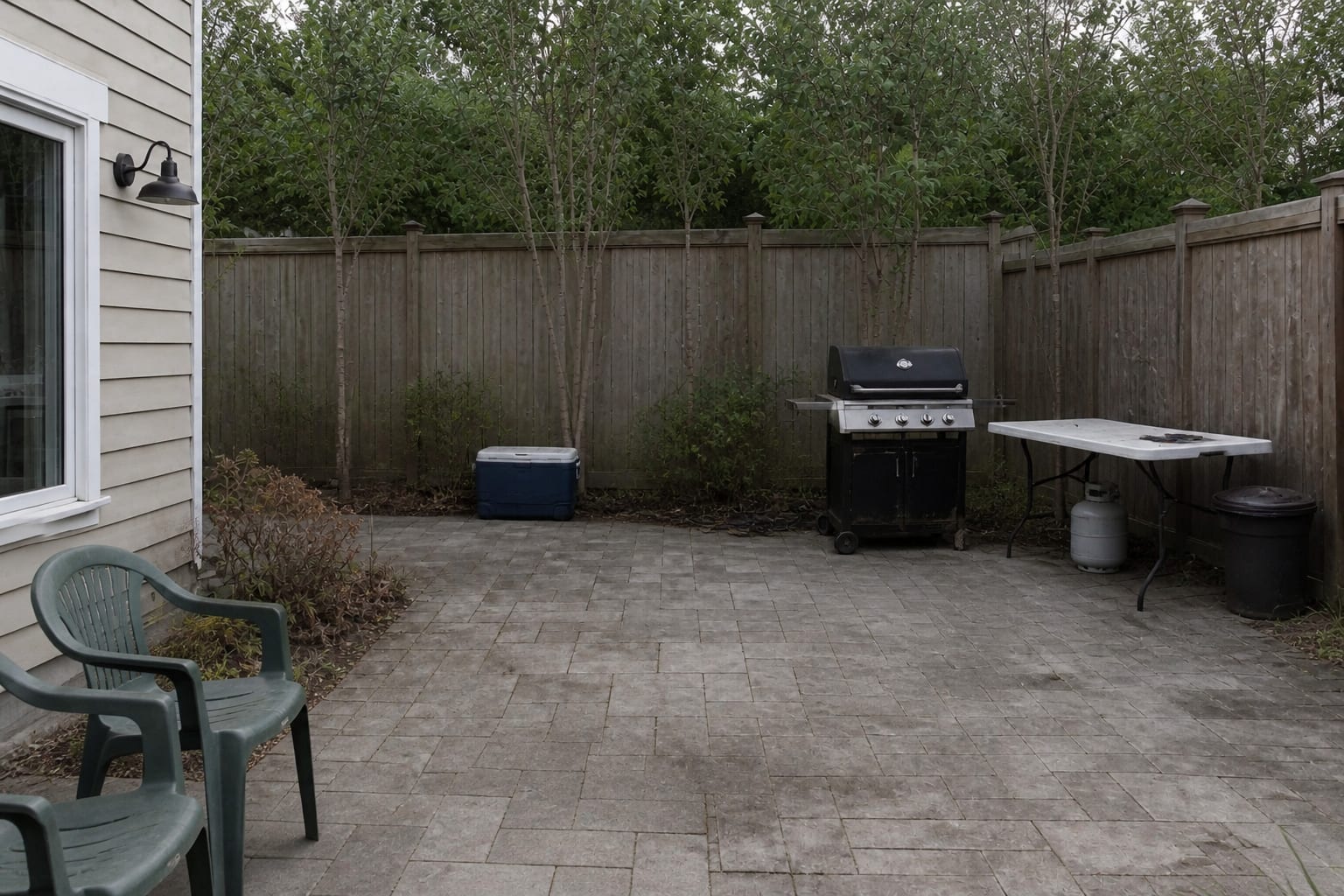 Underplanned patio corner with a freestanding grill, folding table, cooler, scattered tools, and chairs blocking the cooking path.