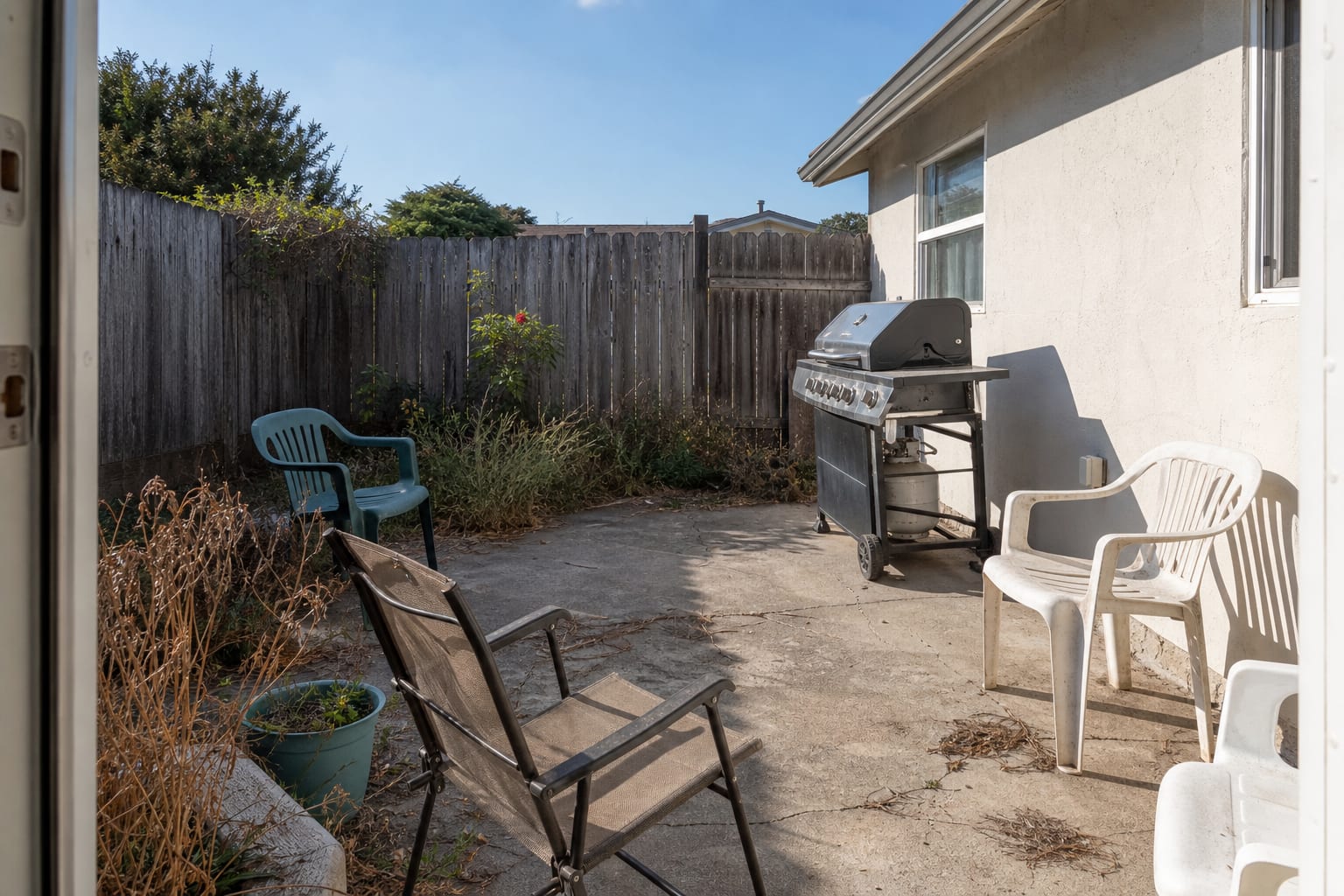 Exposed patio with a freestanding grill, no overhead shade, scattered chairs, and a blank wall behind the cooking area.