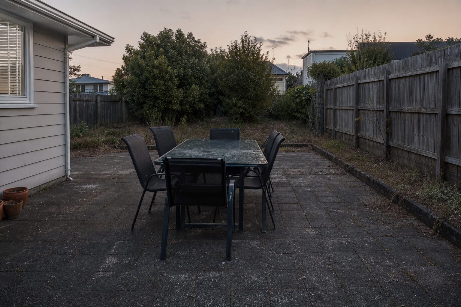 Garden dining table at dusk with dark paving, unlit planting beds, no path markers, and a patio that feels unfinished after sunset.
