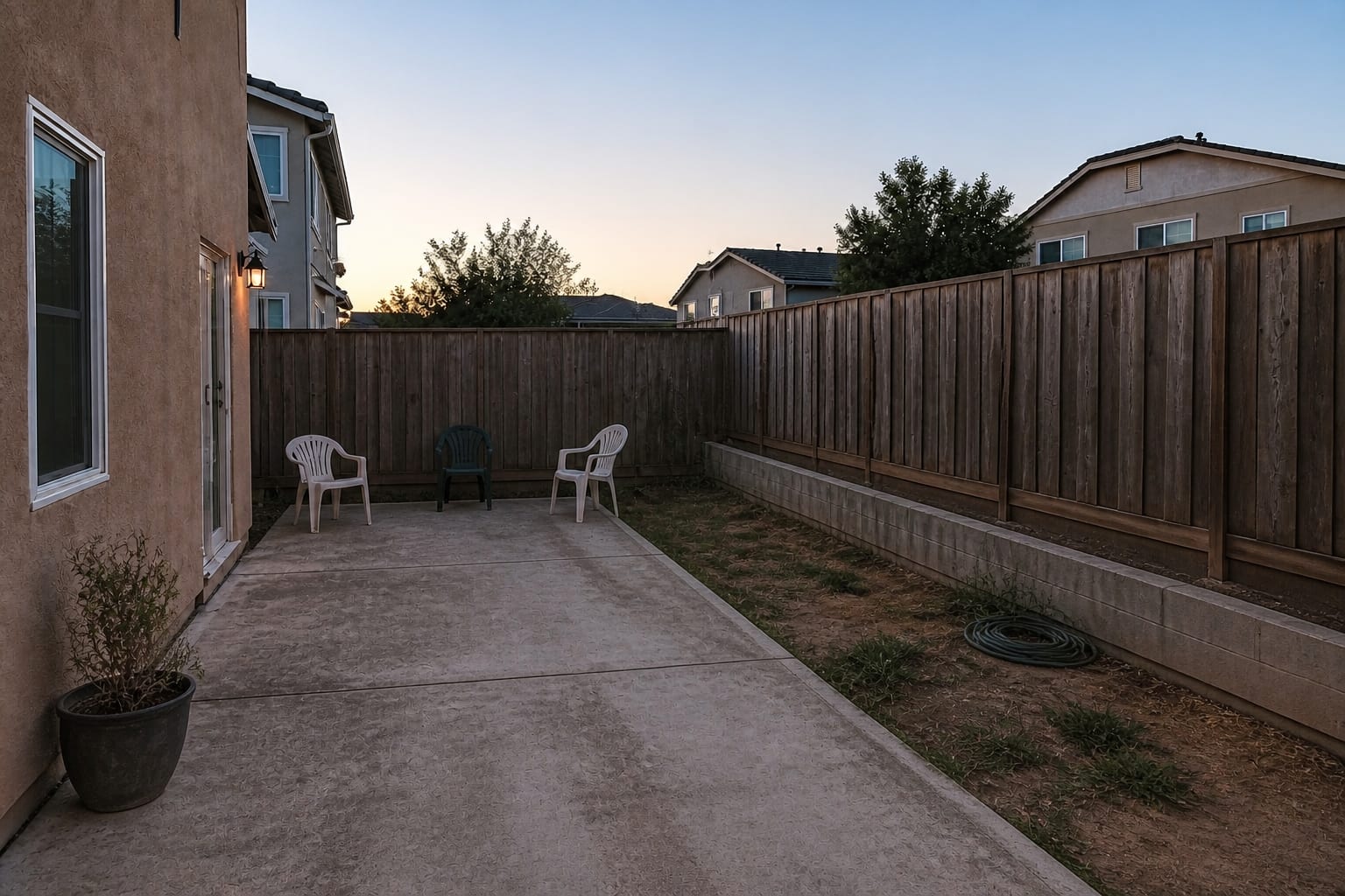 plain concrete patio behind a house with scattered chairs, no shade, no rug, bare fence, and one harsh exterior light.