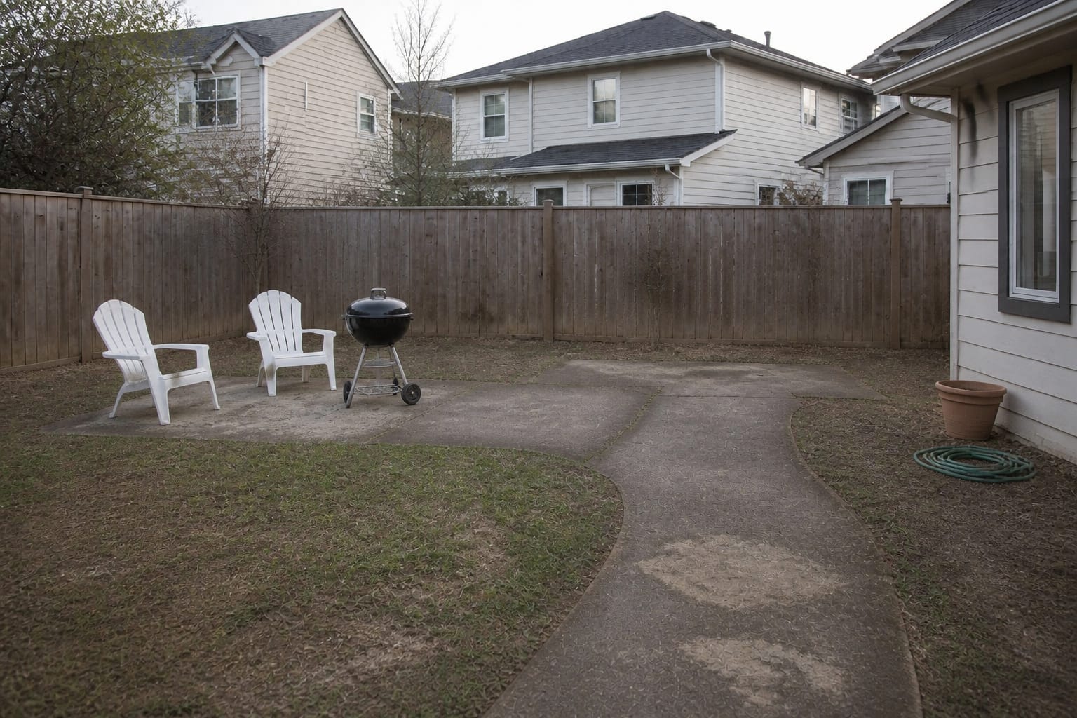 Bare backyard with patchy lawn, a single rolling grill, and plastic chairs before redesign