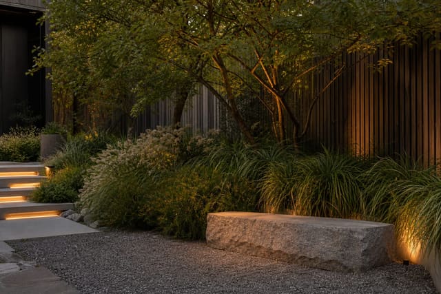 quiet backyard garden corner with low stone seat, layered grasses, small tree canopy, gravel floor, and warm low lighting
