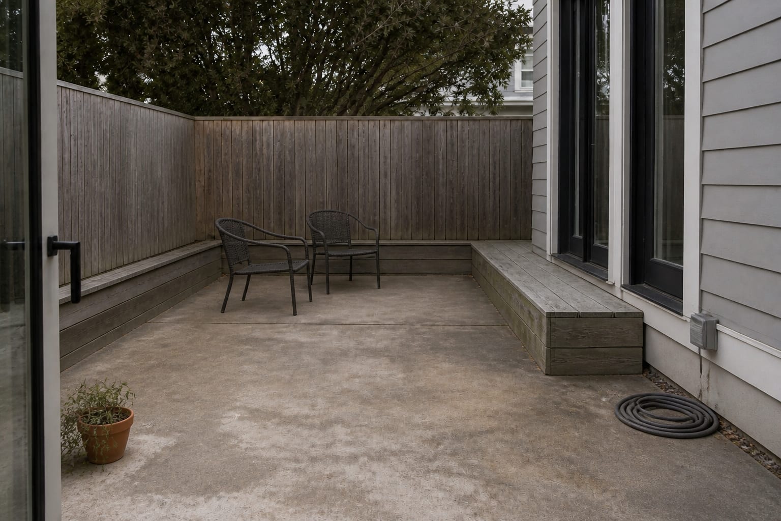 bare concrete patio with two small mismatched pots, exposed fence panels, and outdoor chairs floating without planted edges