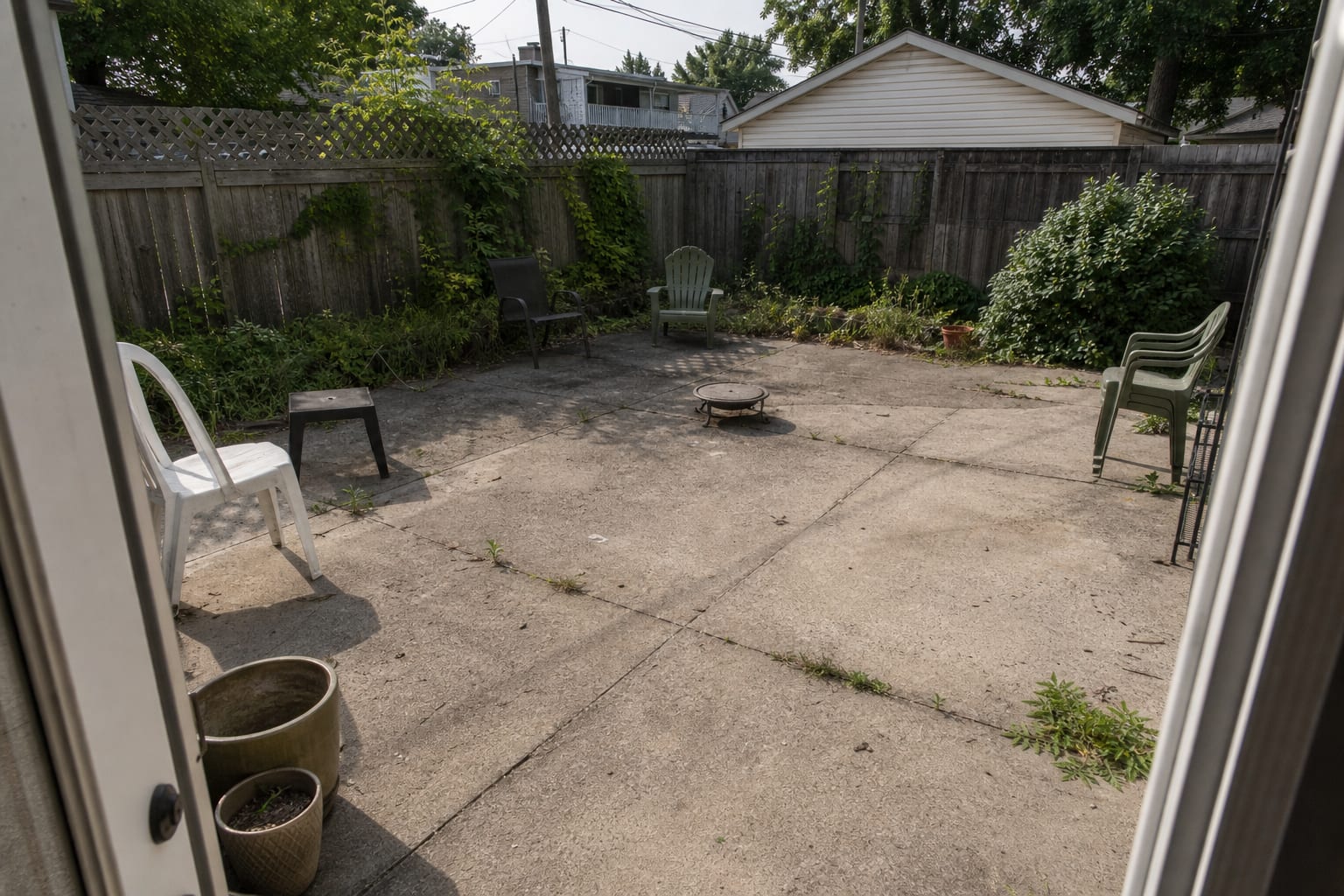 bare concrete patio with two small mismatched pots, exposed fence panels, and outdoor chairs floating without planted edges