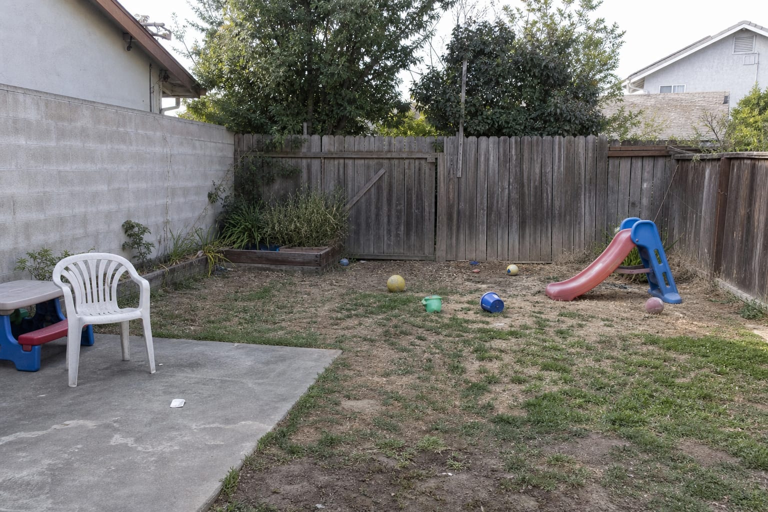 plain backyard corner with patchy grass, a cheap plastic slide, scattered toys, no shade, and no defined play surface