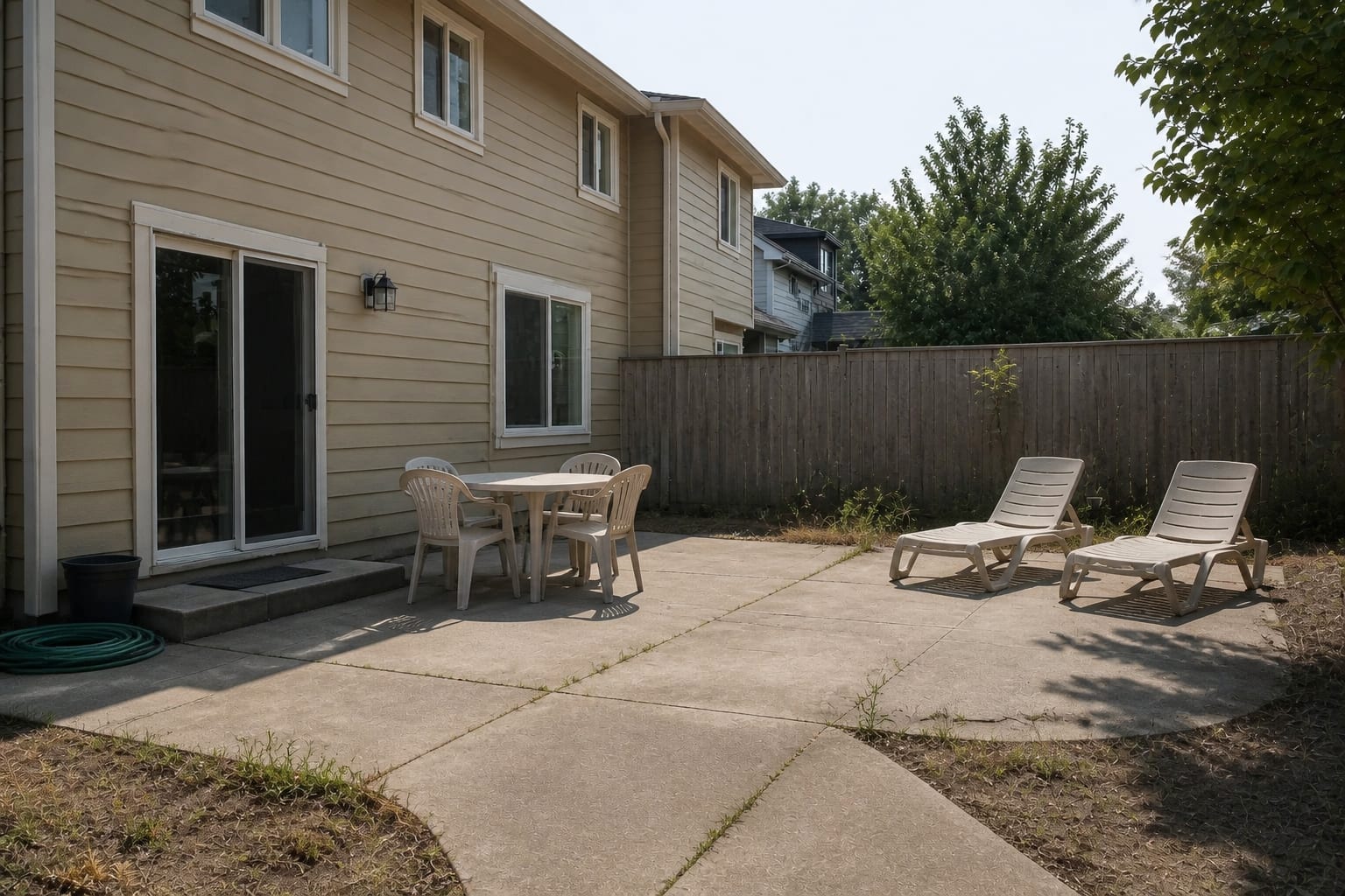 concrete patio with a small table, uncovered lounge chairs, strong afternoon glare, and no shade over the door or seating