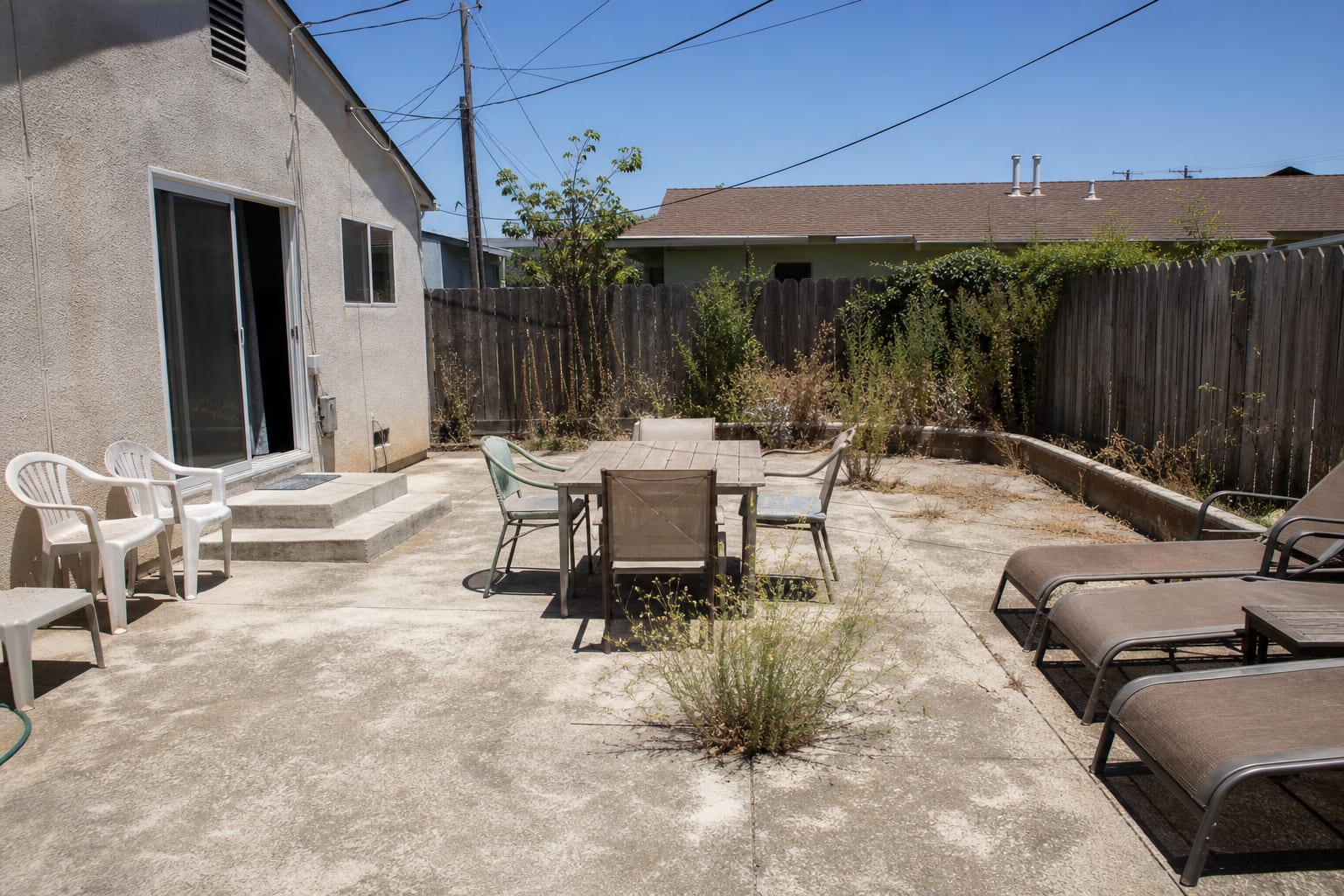 concrete patio with a small table, uncovered lounge chairs, strong afternoon glare, and no shade over the door or seating