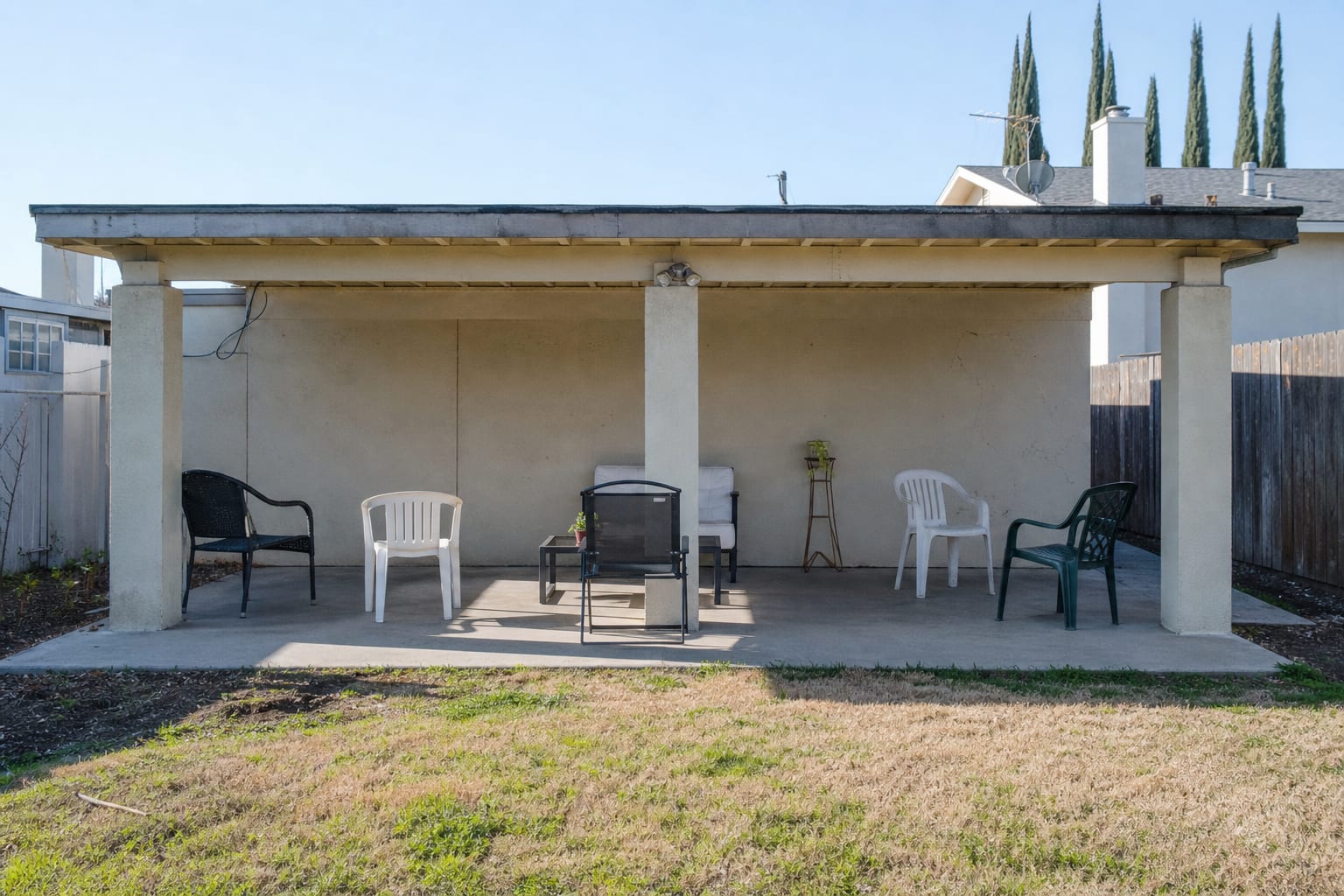 Plain concrete patio with bare square pillars, a flat rear wall, mismatched chairs, and no texture or lighting around the seating area.