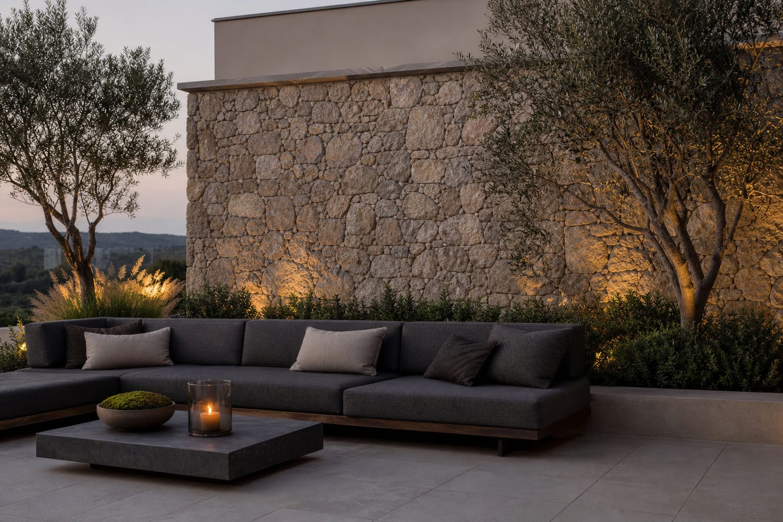 Outdoor stone veneer feature wall with a slim concrete cap, charcoal sofa, olive trees, and a simple porcelain patio floor.