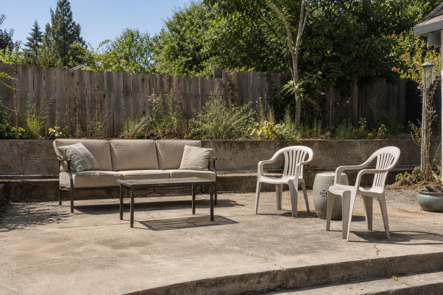 Bare outdoor sofa with thin beige cushions, two undersized faded pillows, and a patio seating area that looks unfinished.