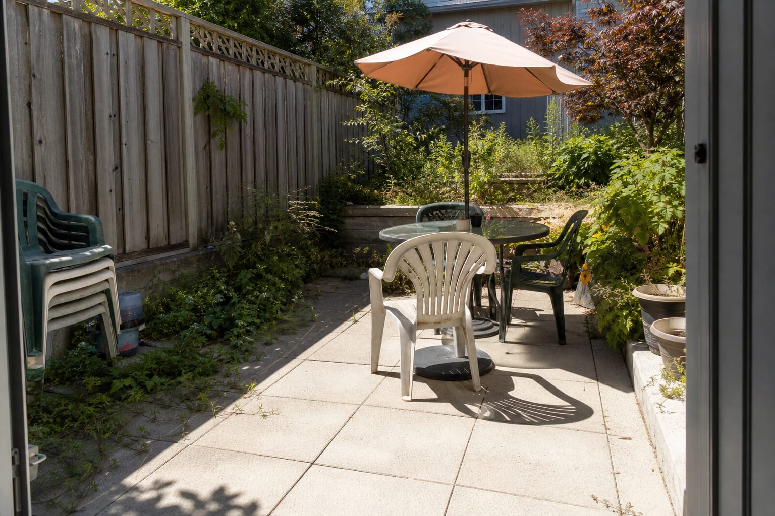 uncovered patio dining set on pale pavers with harsh midday glare, undersized umbrella base, and chairs crowded near the door