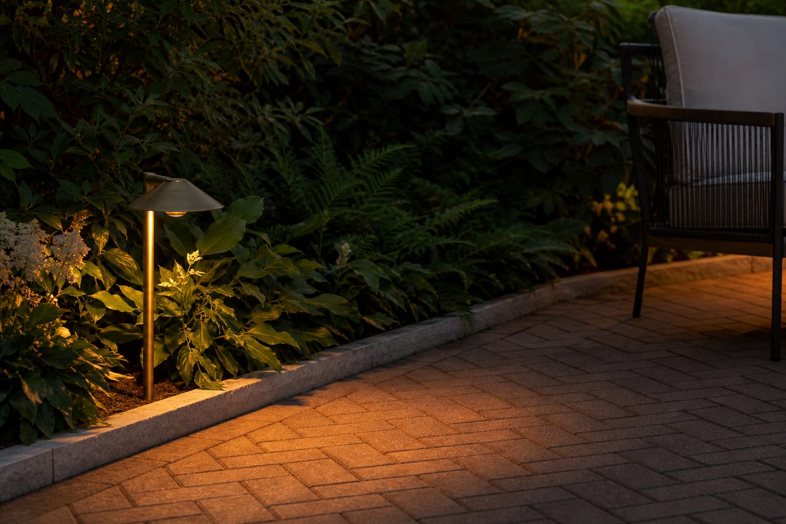 shielded brass path light casting a warm pool across brick pavers with dark planting behind the walkway