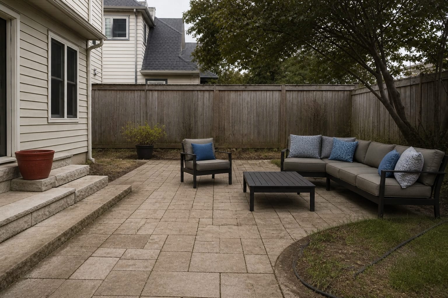 small backyard patio with gray cushions, blue pillows, red pots, beige pavers, and no clear outdoor color palette
