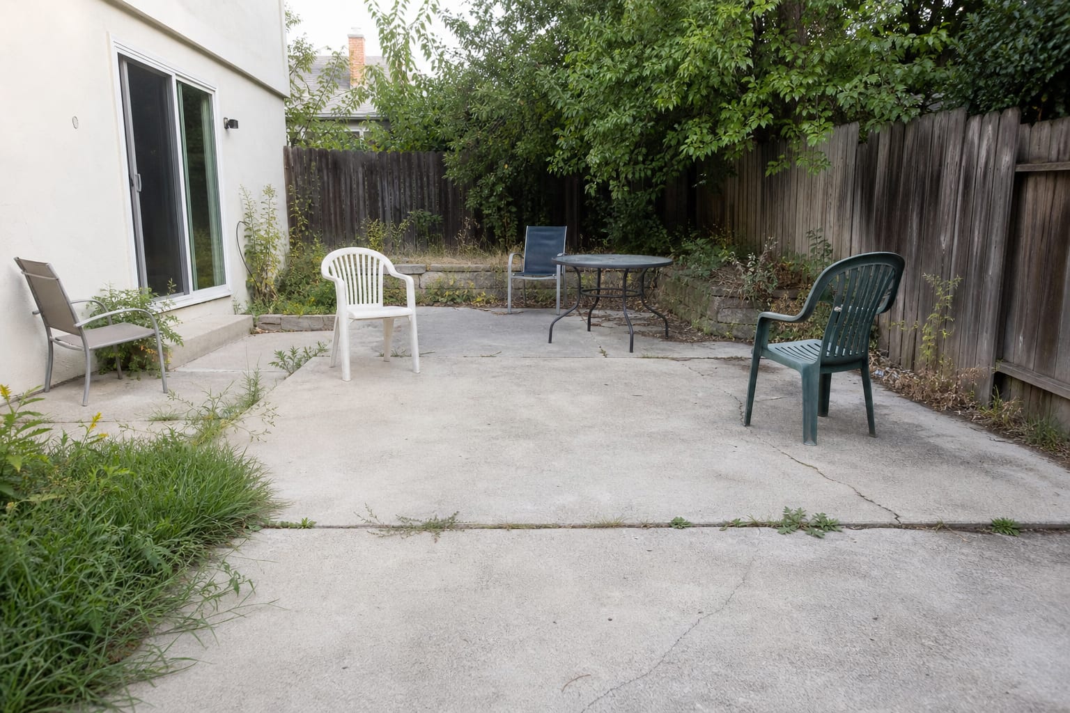 cracked concrete patio outside a back door with mismatched chairs, exposed edges, weeds in joints, and no clear outdoor room layout.