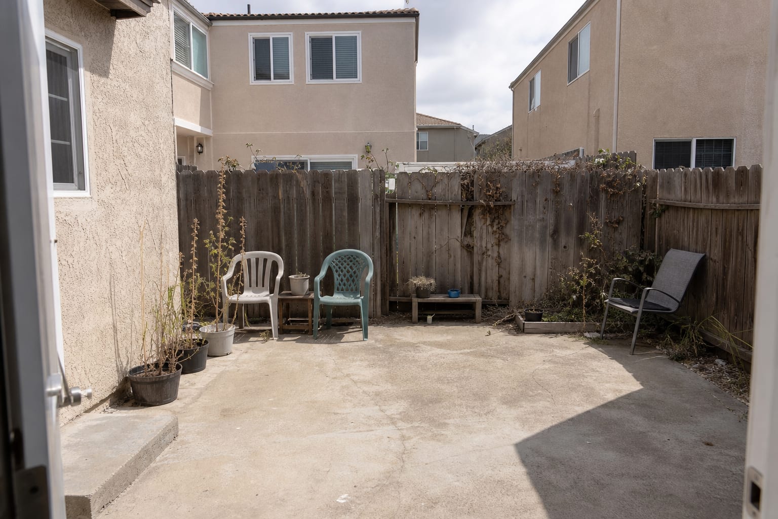 Exposed patio with low furniture, open fence gaps, bare concrete, and direct views from nearby neighbor windows.