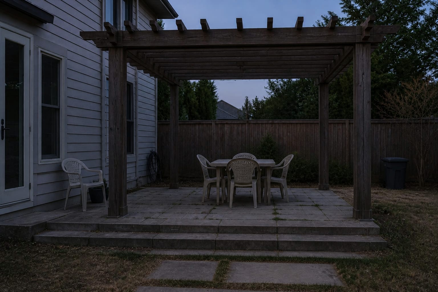 dark backyard pergola over a patio table with no visible lighting, dim paving, and chairs disappearing into evening shadow.