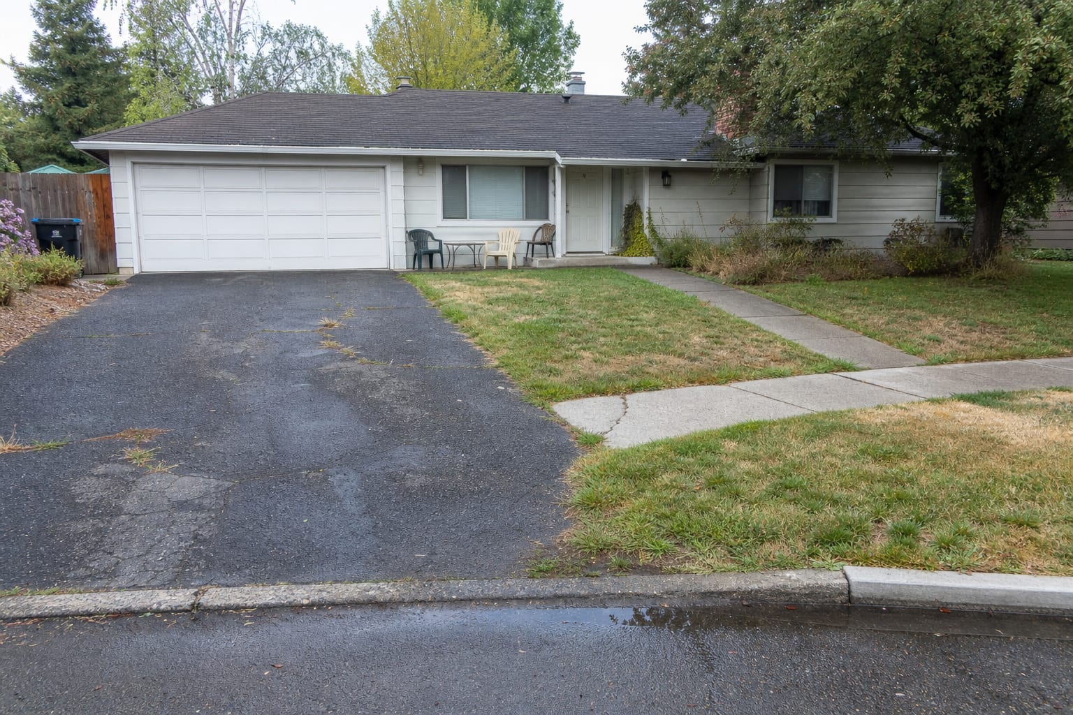cracked asphalt driveway beside a plain front lawn with water collecting near the street after rain
