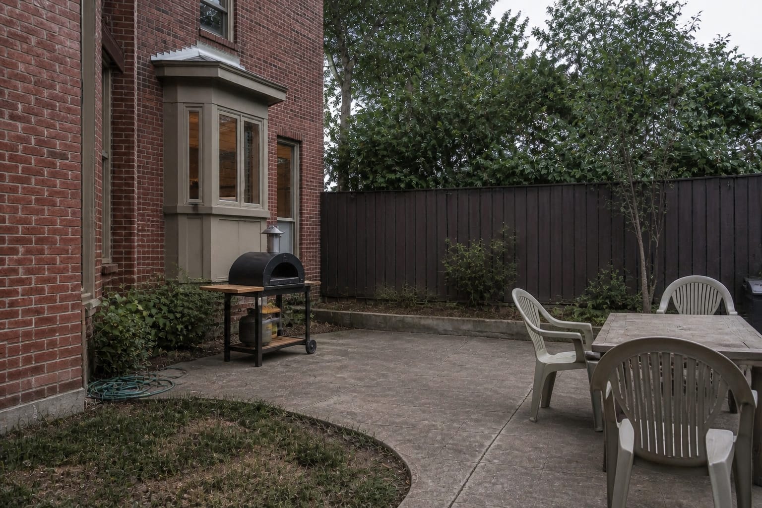 plain concrete patio corner with a portable pizza oven on a small cart, scattered chairs, and no prep surface or lighting.