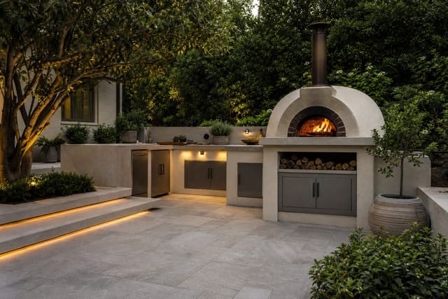 wood fired pizza oven on a porcelain tile patio with a prep counter, herb planters, and warm step lighting