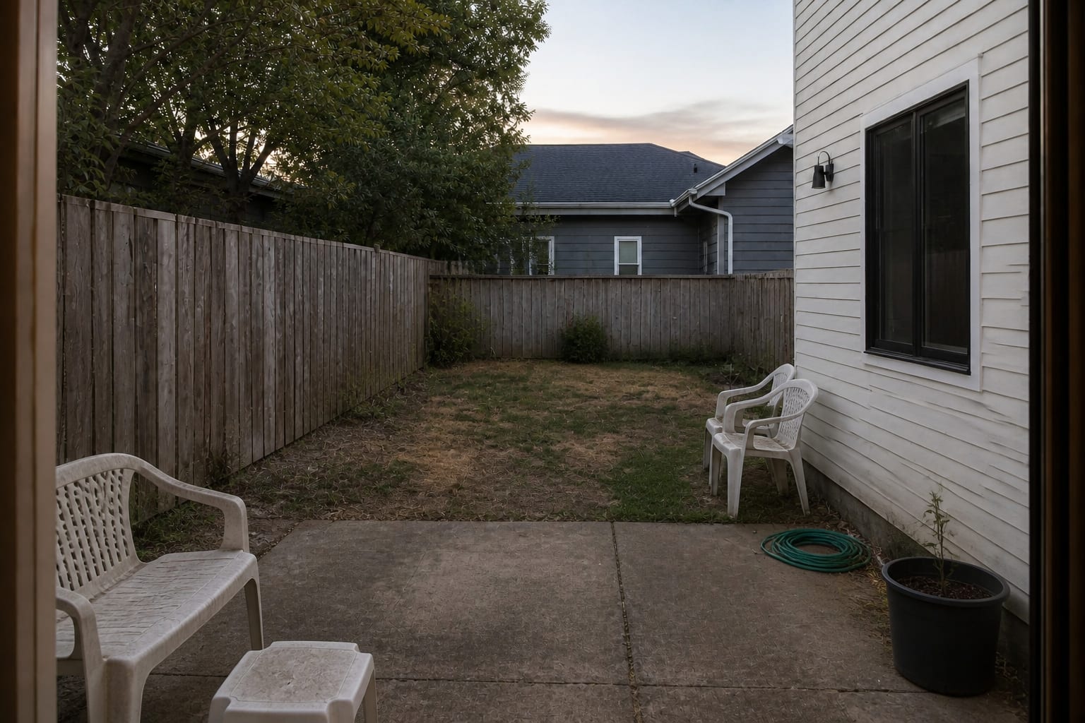 Narrow grass backyard with a blank fence, scattered chairs, no shade, and no defined place to cool off.