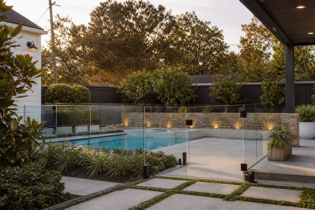 glass and aluminum pool fencing around a compact backyard pool with planted edges, stone paving, and a clear self-closing gate