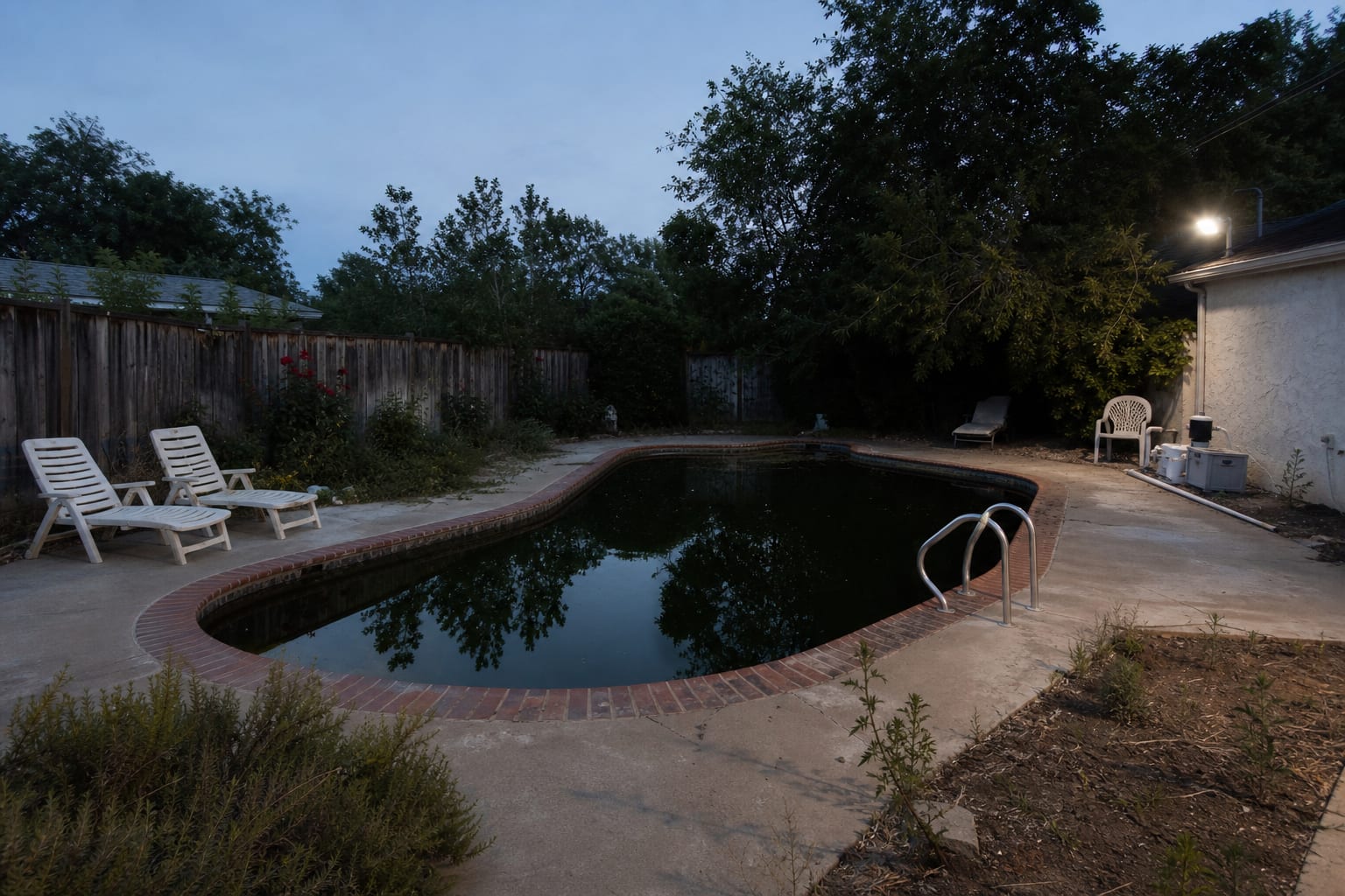 dark backyard pool area with black water, harsh wall floodlight, unmarked coping edge, and scattered furniture near the deep end