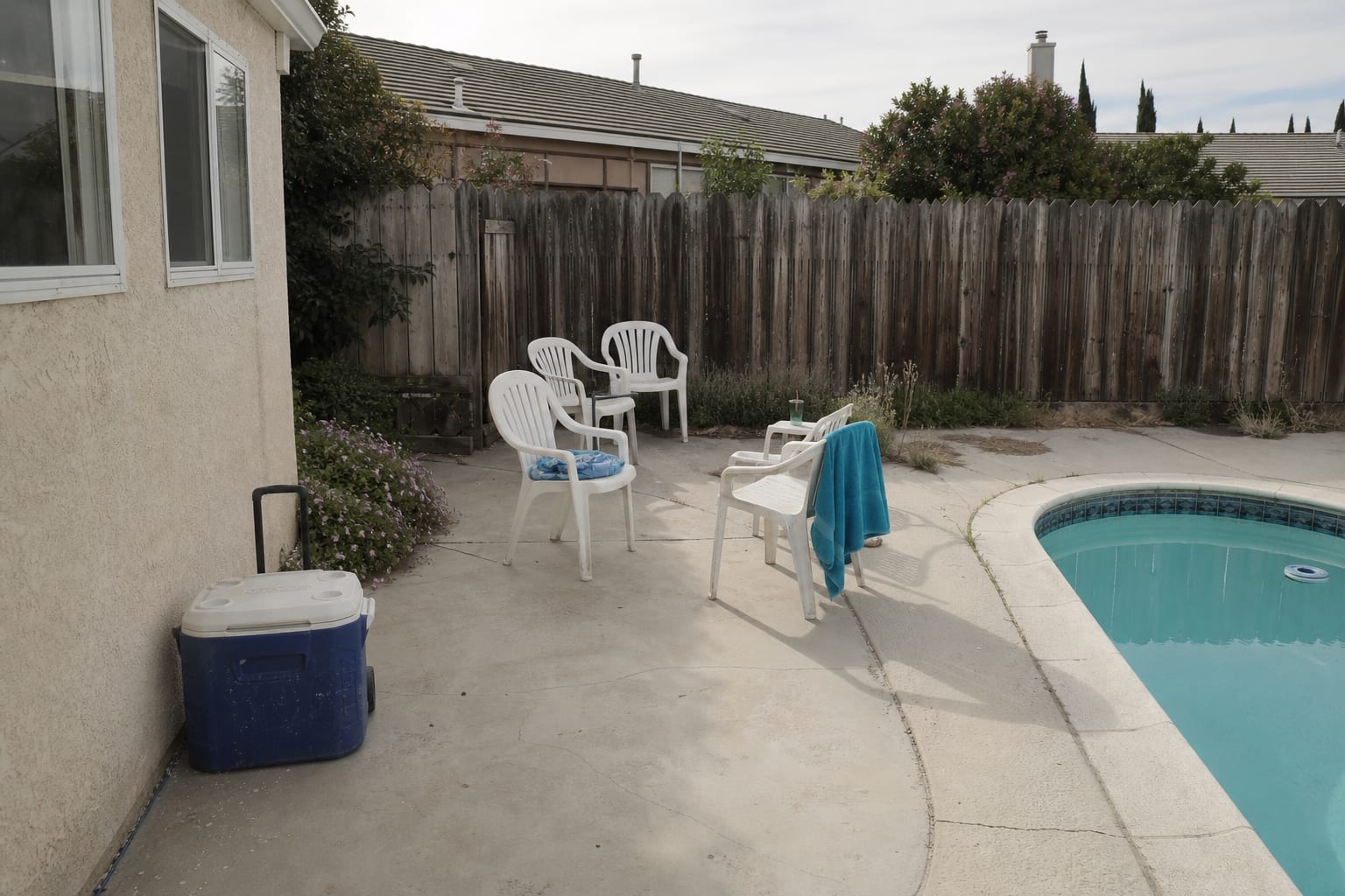 pool patio corner with a plastic cooler, scattered towels, no shade, and lounge chairs blocking the walking path around the water.