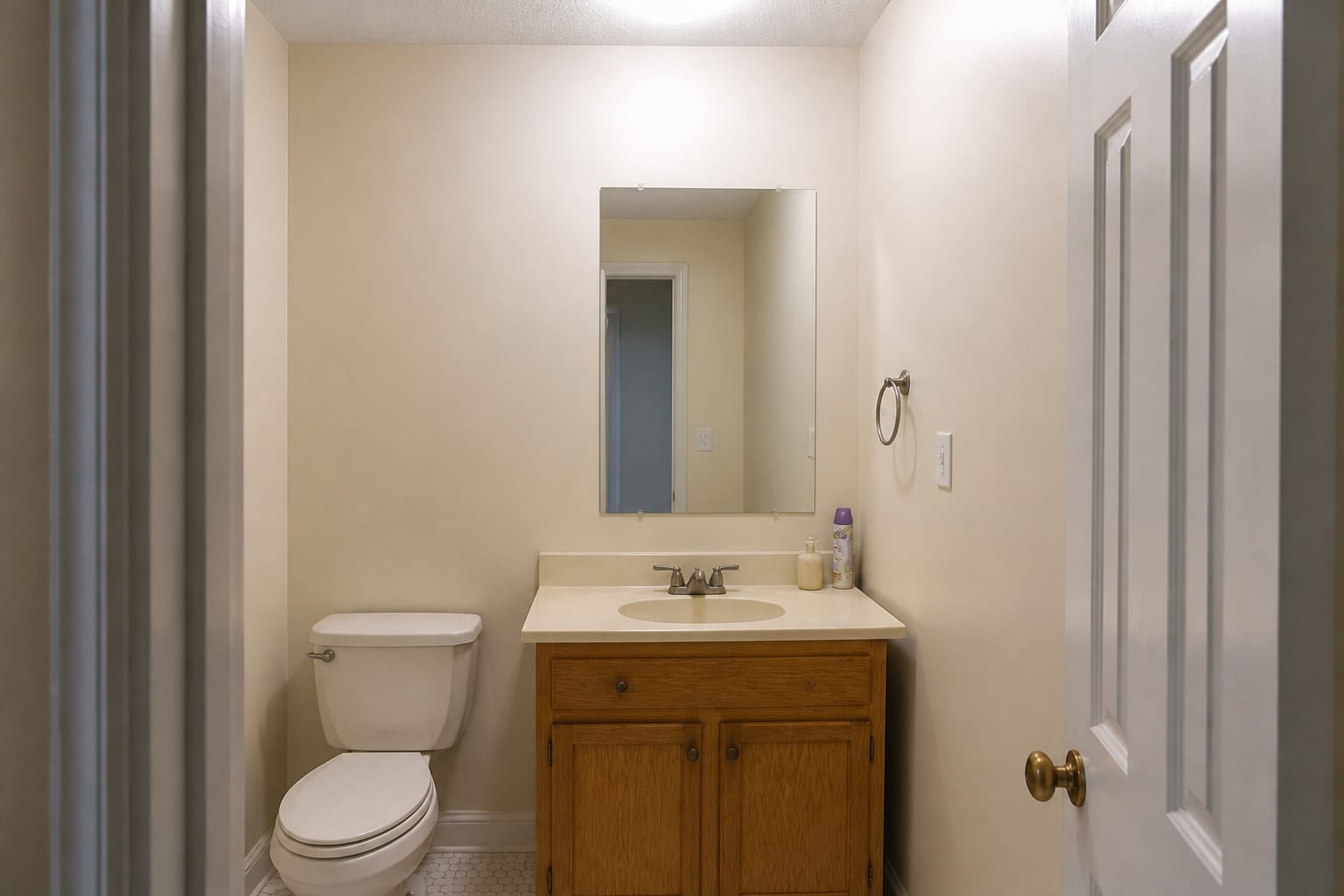 Plain powder room with beige walls, builder mirror, small vanity, and cold overhead light.