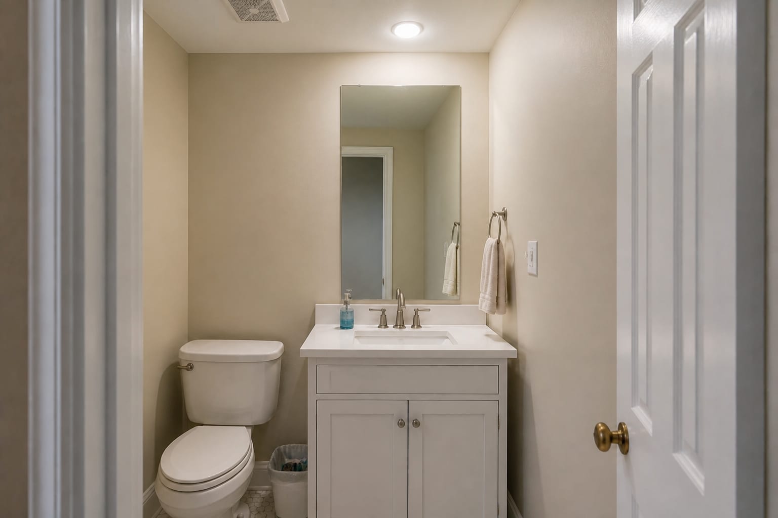 Plain powder room with beige walls, builder mirror, small vanity, and cold overhead light.