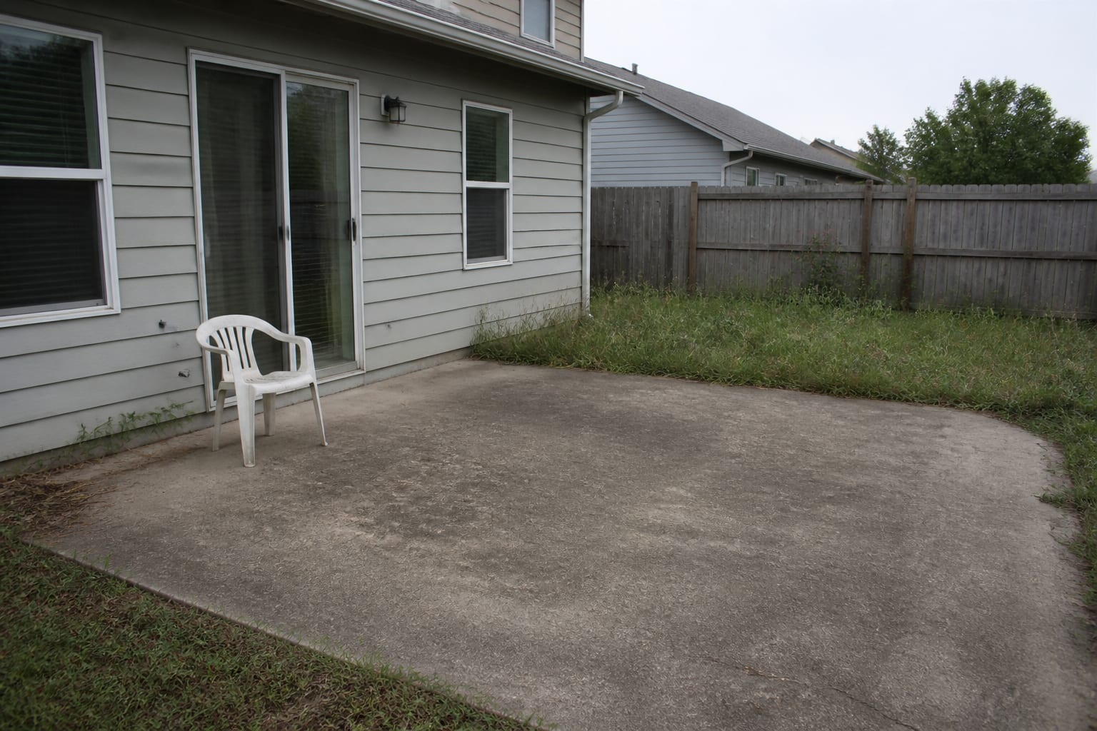 A plain concrete patio with mismatched plastic furniture, overgrown edges, and no defined seating area
