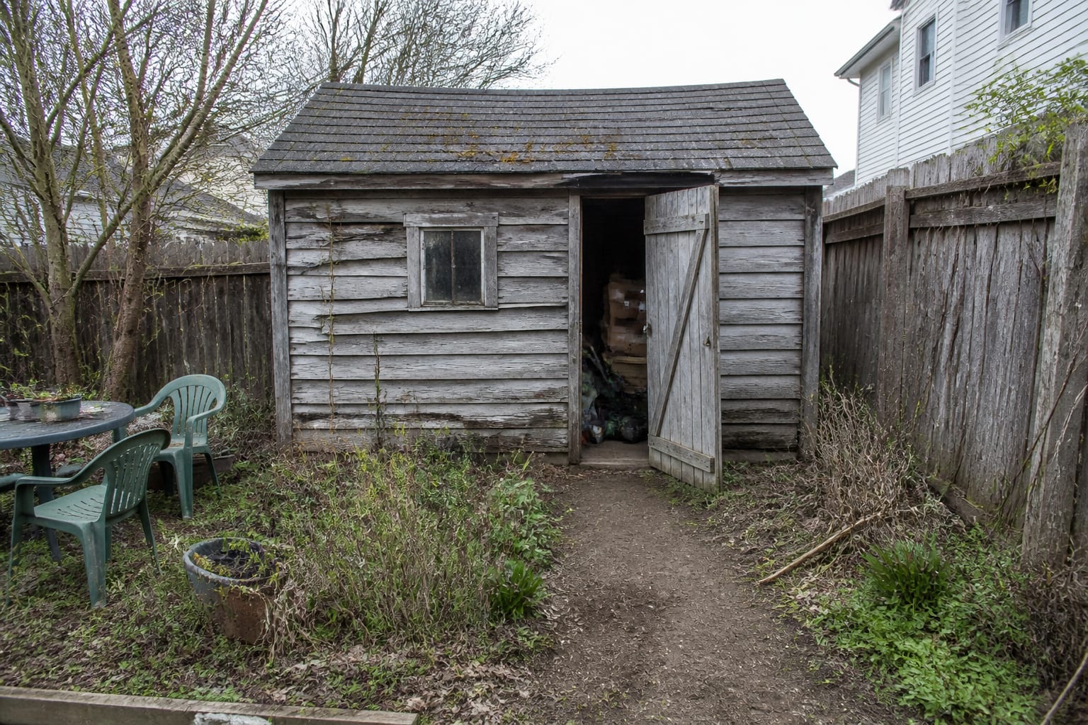 A neglected backyard wooden shed with weathered gray siding, a small dark window, cluttered interior, and a bare dirt path before any redesign.