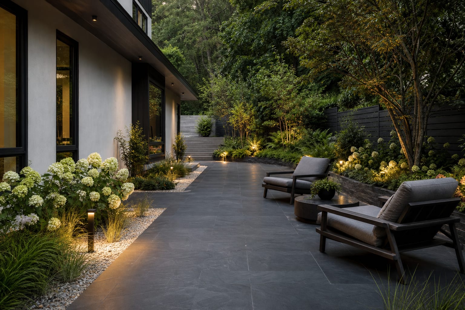 dark slate patio with low lounge chairs, pale gravel planting beds, and warm path lights beside a simple modern home