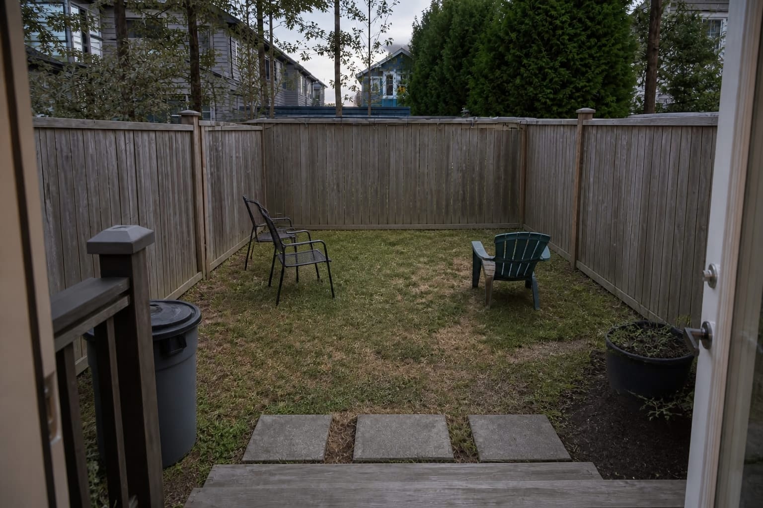 Narrow backyard with patchy grass, random chairs, a bare fence, and no clear seating zone or planted edge.