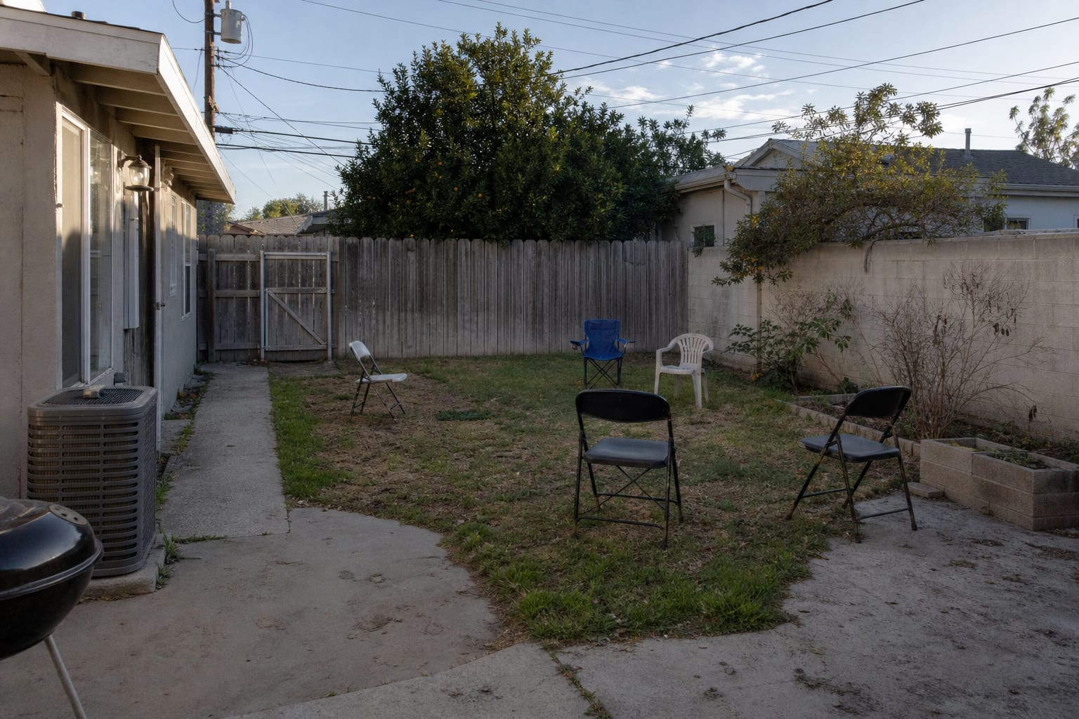 Narrow backyard with patchy grass, random chairs, a bare fence, and no clear seating zone or planted edge.