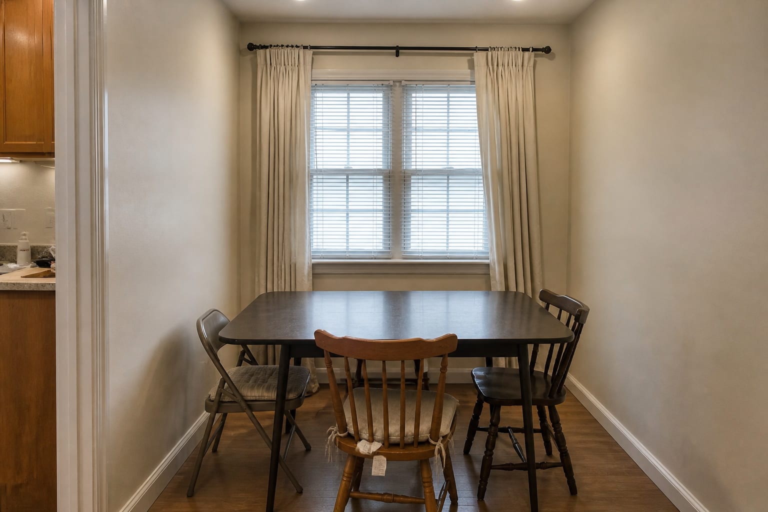 Cramped breakfast nook with an oversized rectangular table, mismatched chairs, heavy drapes, and harsh overhead can lights.