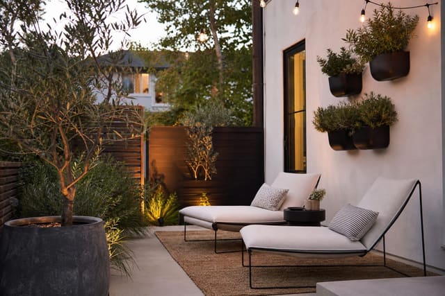 small patio with slim lounge chairs, potted olive tree, woven rug, wall planters, and warm string lights at dusk