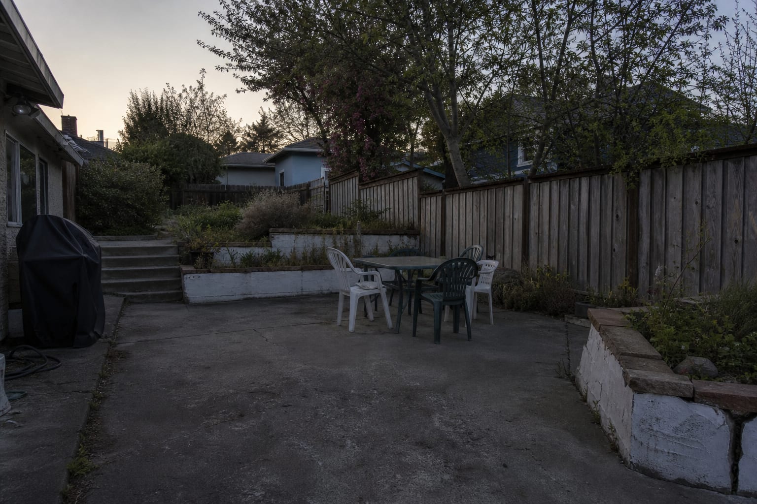 Backyard patio at dusk with dark paving, unlit fence line, shadowy steps, and a dining table that disappears into the yard.