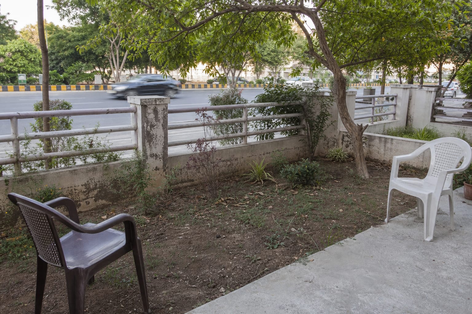 under designed road-facing garden with a low open fence, exposed chairs, bare soil, and visible traffic beyond the boundary