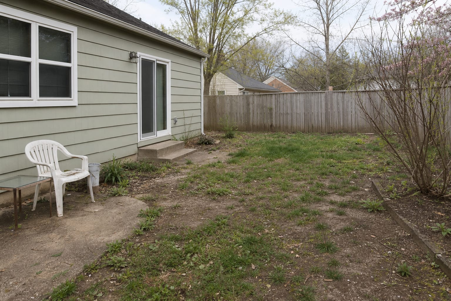 patchy dirt and thin grass beside a garden bed with no clear patio shape, no seating zone, and no stable surface for chairs.