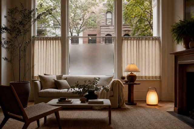 street-facing living room window with frosted lower glass, linen café curtains, plants, and warm lamp light