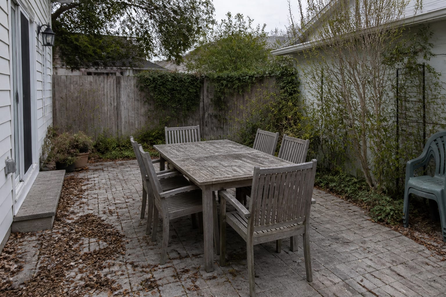 A neglected teak patio dining set weathered to a dull, blotchy grey with bare cushion-less chairs on a dirty patio