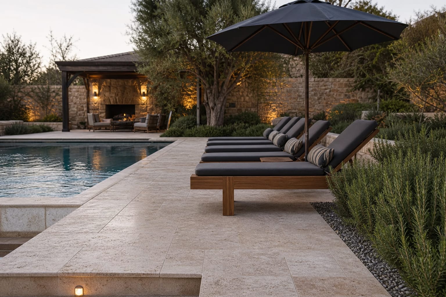 travertine pool deck with bullnose coping, charcoal umbrella, teak loungers, and rosemary planted along a gravel edge
