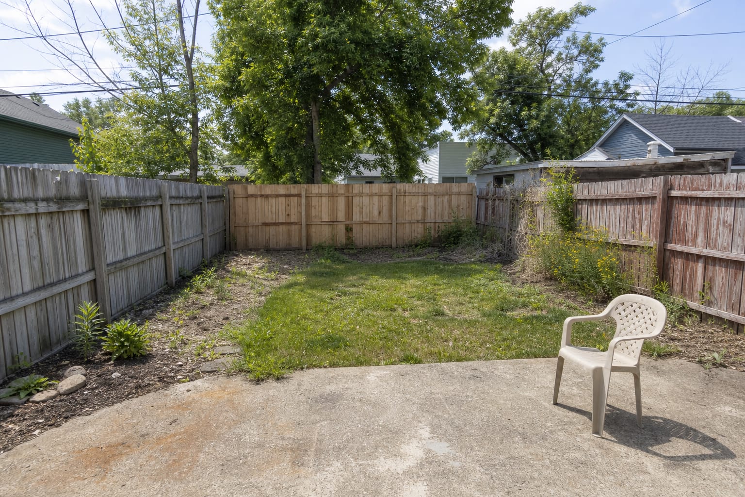 Narrow fenced backyard with patchy grass, exposed soil, one small patio chair, and no layered planting or shade structure.