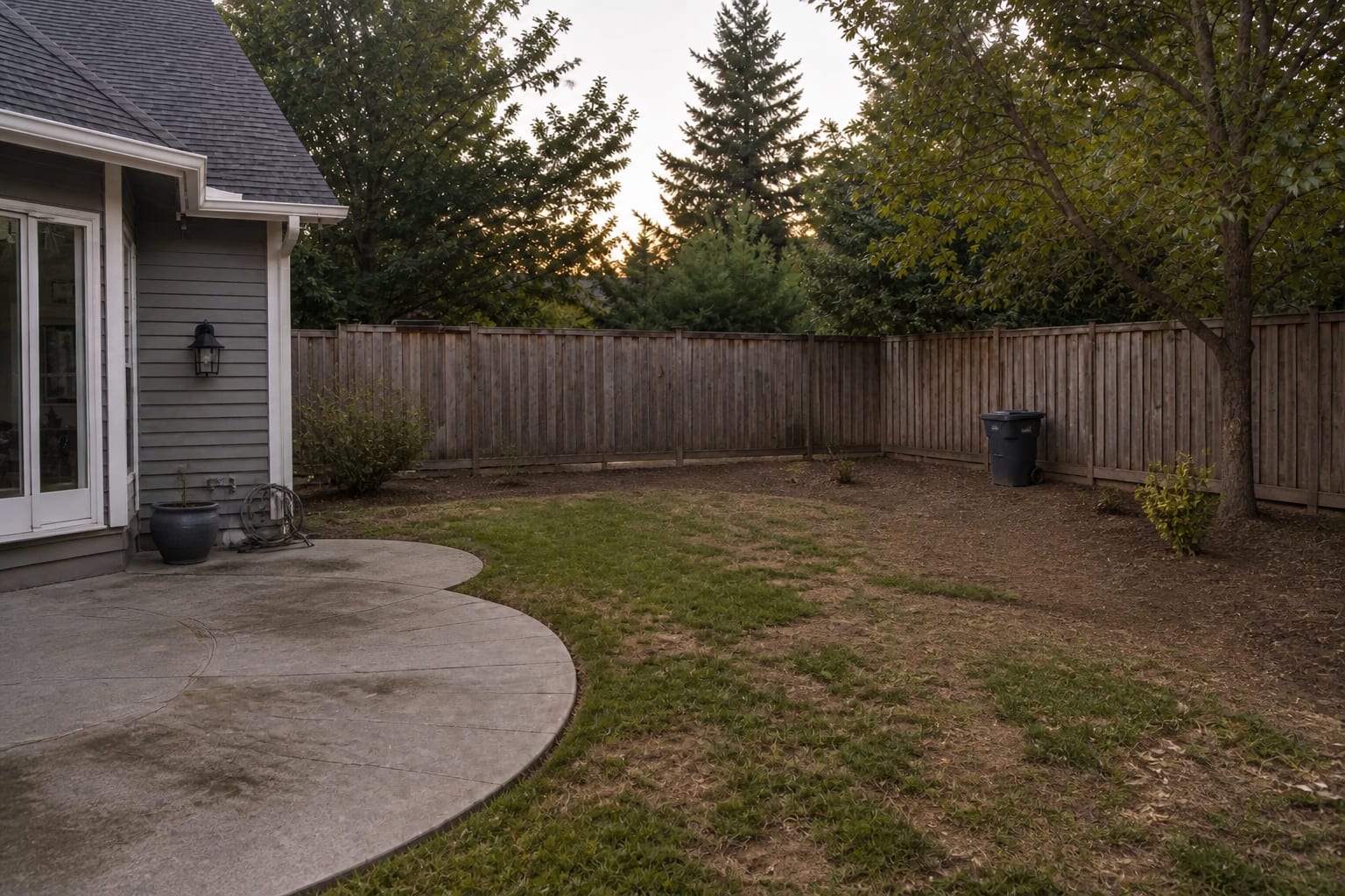 Patchy backyard grass beside a plain patio with no path, no planting structure, and bare soil near the fence.