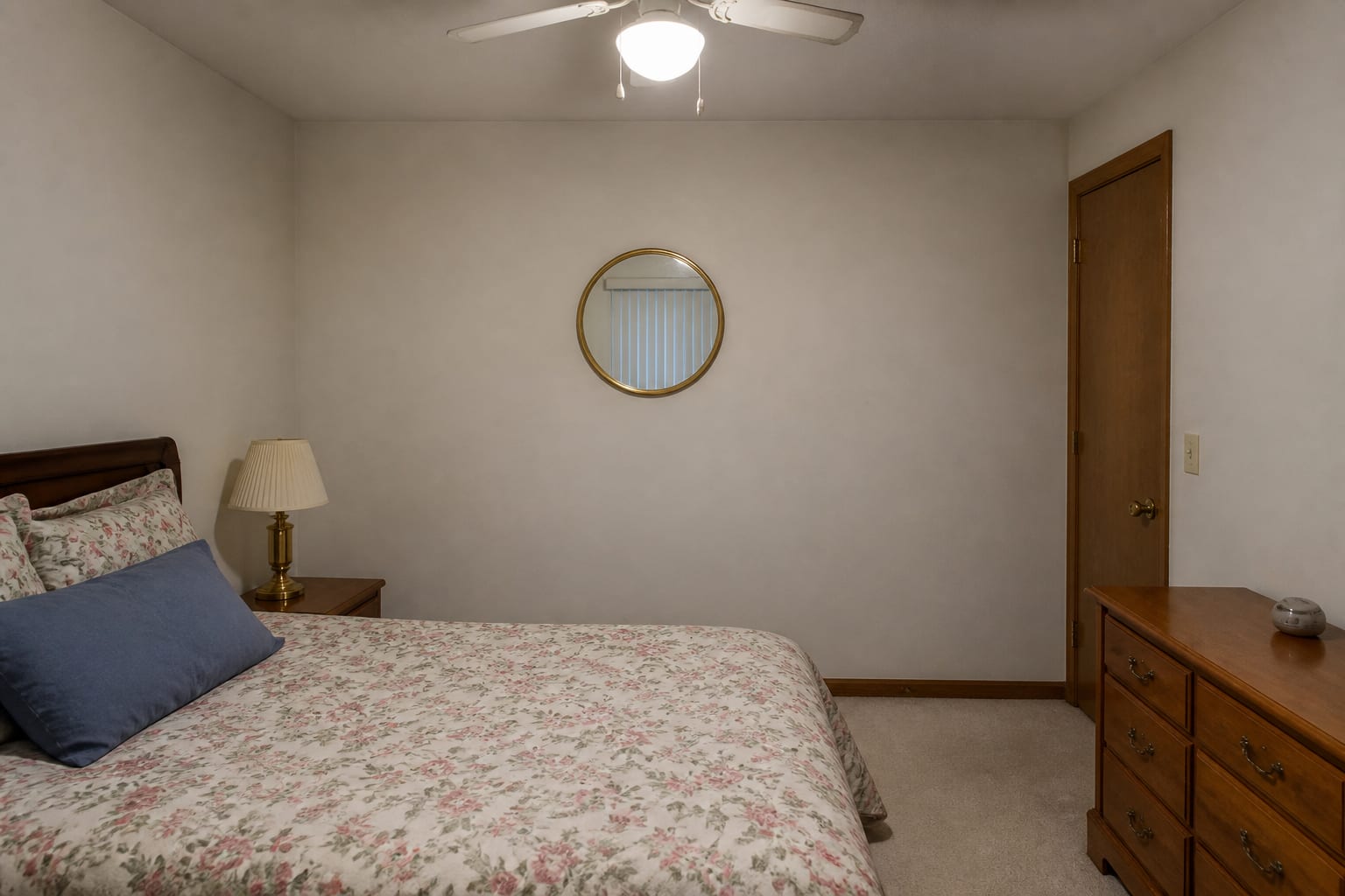 Windowless bedroom with deep plum walls, a small round mirror away from the light, floral bedding, and a cold ceiling-fan light.